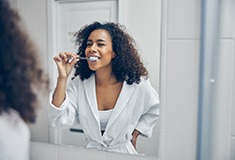 Woman in robe smiling while brushing her teeth in bathroom